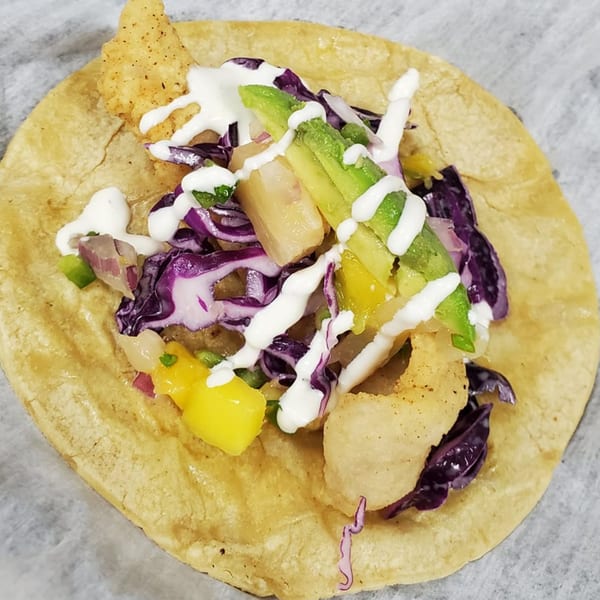 Close-up of a Papas Bar fish taco with fried fish, avocado, red cabbage, mango, and crema.